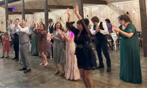 Guests in formal attire dancing and clapping at a rustic barn wedding reception.