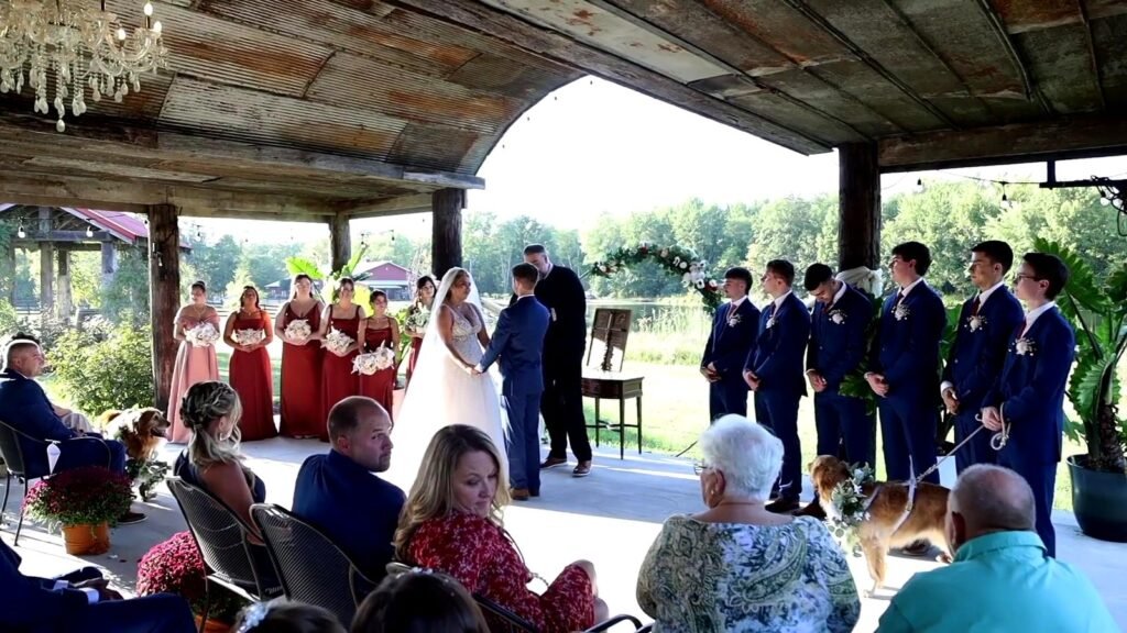 A bride and groom hold hands during their wedding ceremony under a rustic pavilion, surrounded by their wedding party and guests