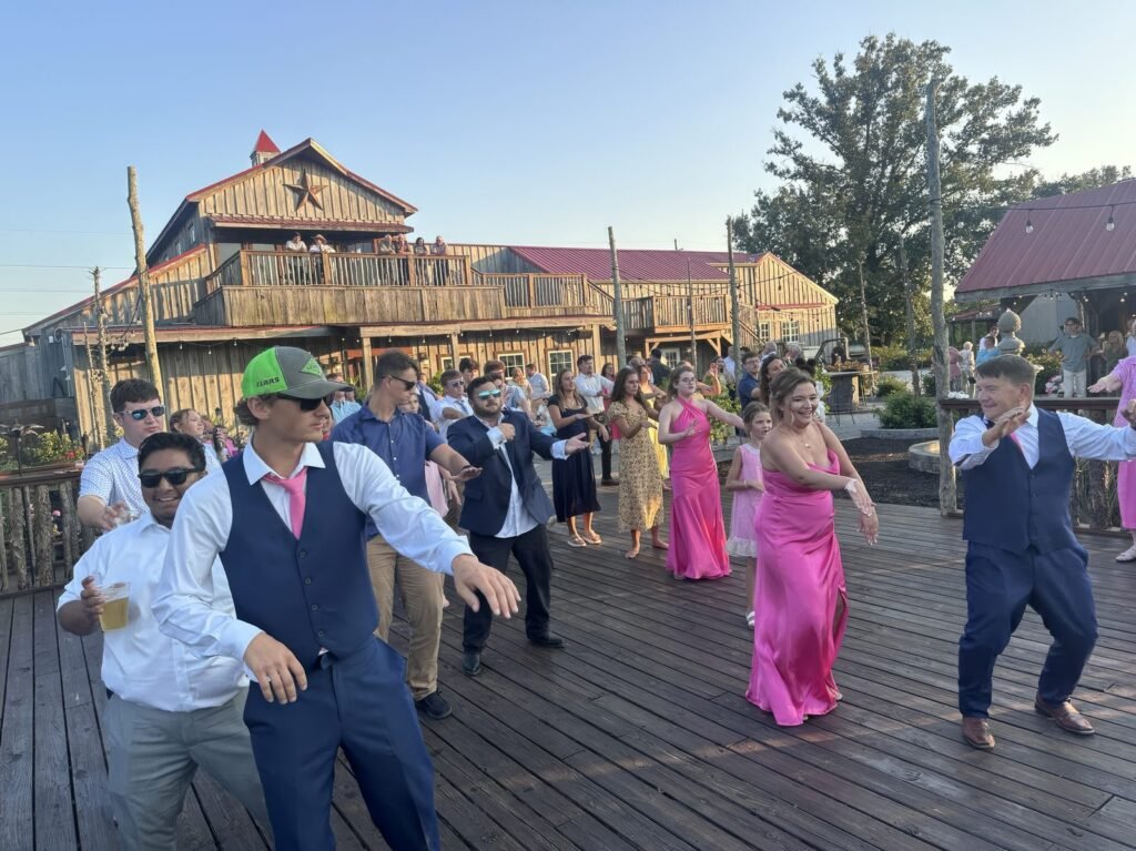 A group of people dancing on an outdoor wooden deck during a sunny wedding reception.