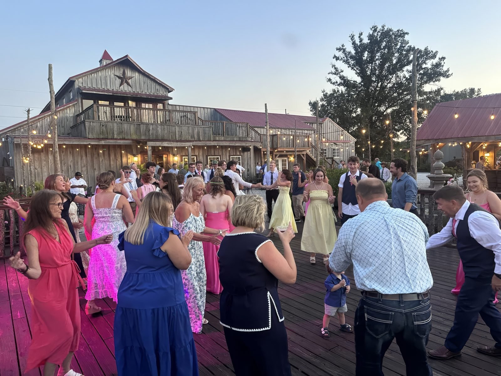 Guests in semi-formal attire dancing on a wooden deck in front of a rustic barn venue under string lights at dusk