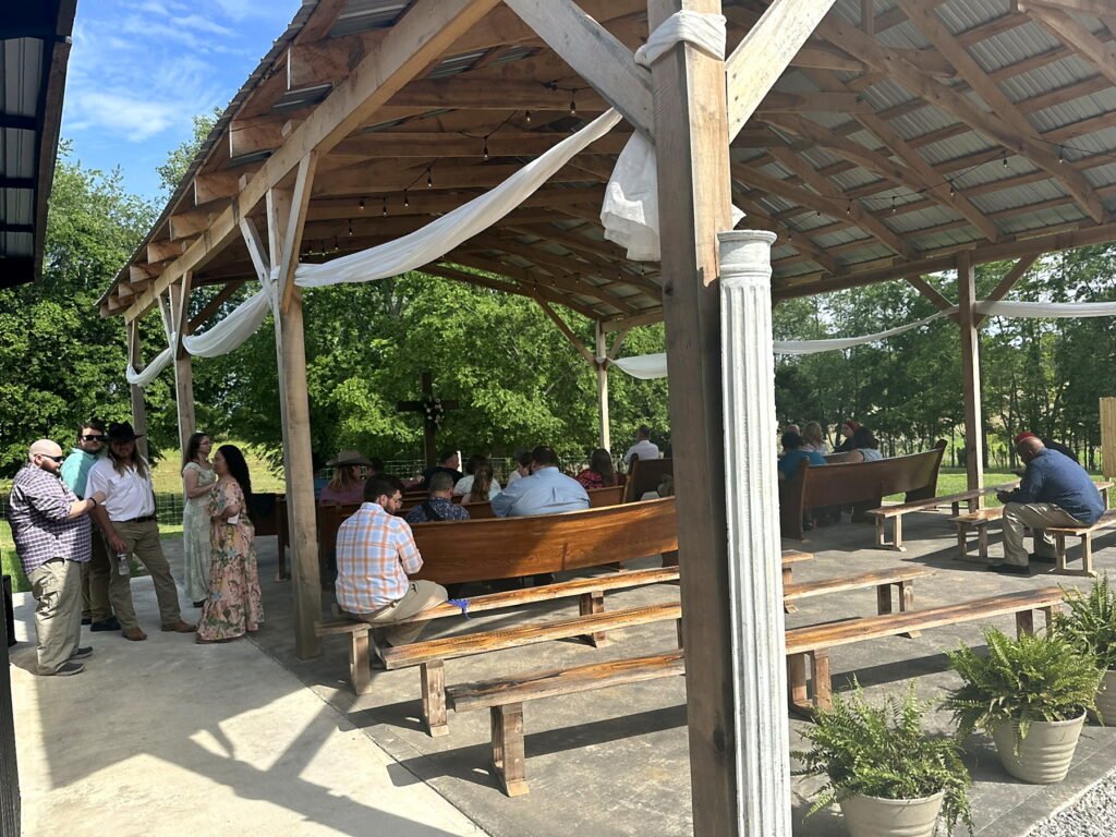 Princeton Kentucky Wedding DJ 9 Guests gather in an open-air wooden pavilion with pews and white fabric draping, with a wooden cross in the background