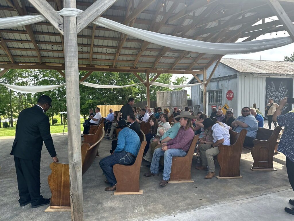 Princeton Kentucky Wedding DJ 8 A man in a black suit speaks from a wooden podium to a seated audience in an open-air wooden pavilion