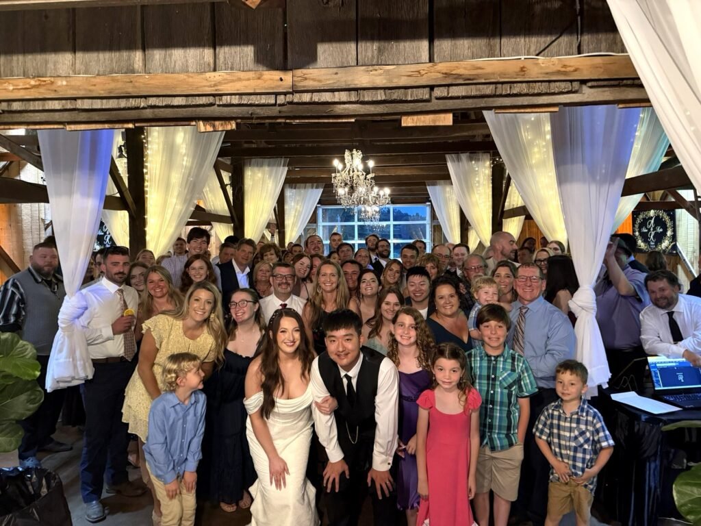 Bride and groom with a large group of guests in the Farmer & Frenchman barn under white drapes and a chandelier