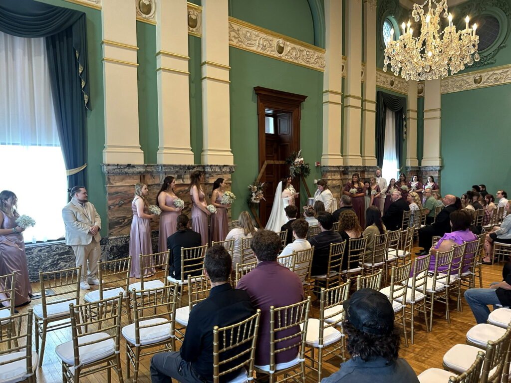 Wedding ceremony in an ornate hall with green walls and gold trim, featuring a bride, bridesmaids in mauve, and seated guests