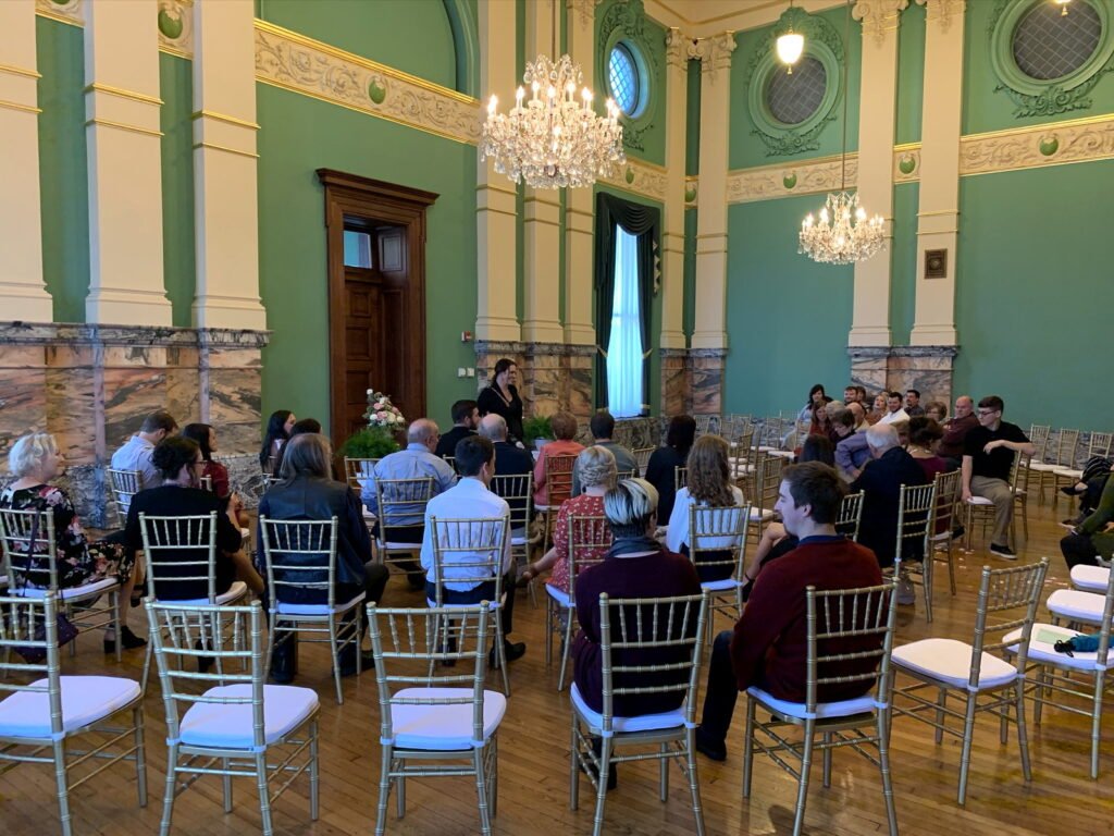 Guests seated in gold chairs in a grand room with green walls and chandeliers, facing a speaker at the front