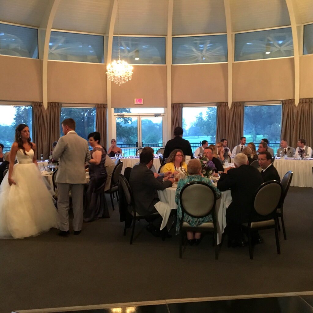 A bride and groom stand near guests seated at tables in a large, bright wedding reception hall