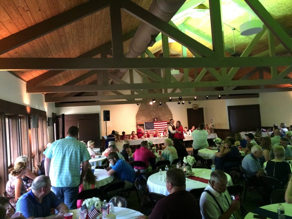 Guests at round tables in a rustic hall with a high beamed ceiling, with a couple speaking at a head table