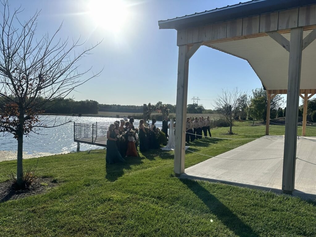 A bride, groom, and wedding party stand before a floral arch for an outdoor wedding ceremony beside a lake
