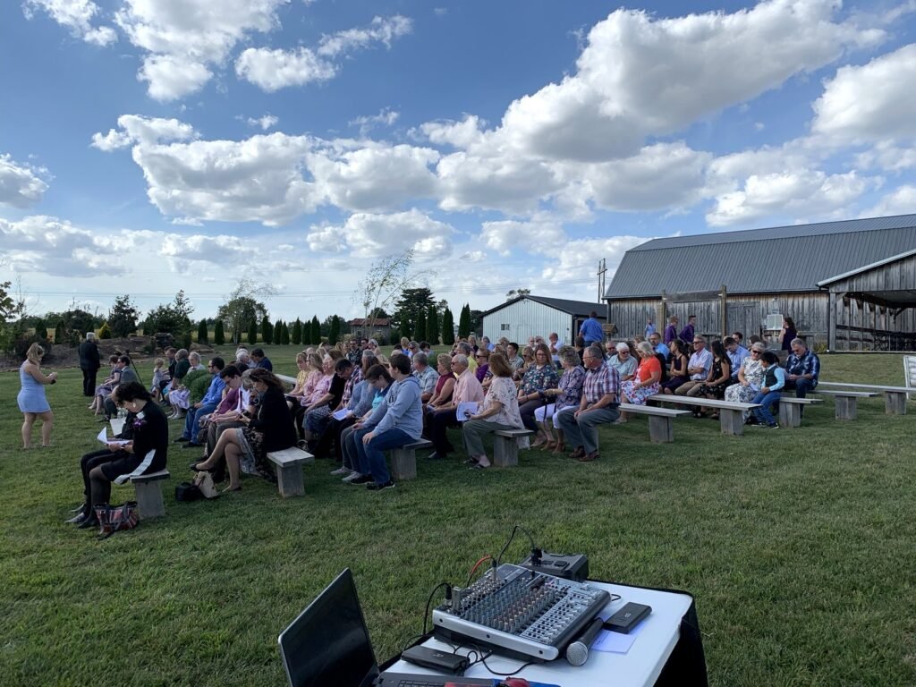 Guests sit on wooden benches in a grassy field for an outdoor event, with a barn in the background and audio equipment in the foreground