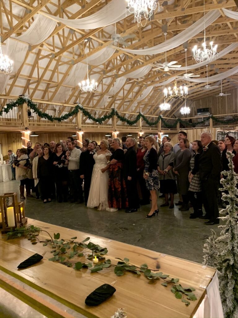 A bride and groom stand with their wedding guests in a rustic barn decorated with white fabric and chandeliers