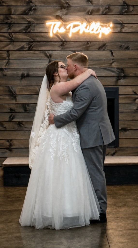Bride and groom kissing in front of a wood wall with a neon sign that reads "The Phillips