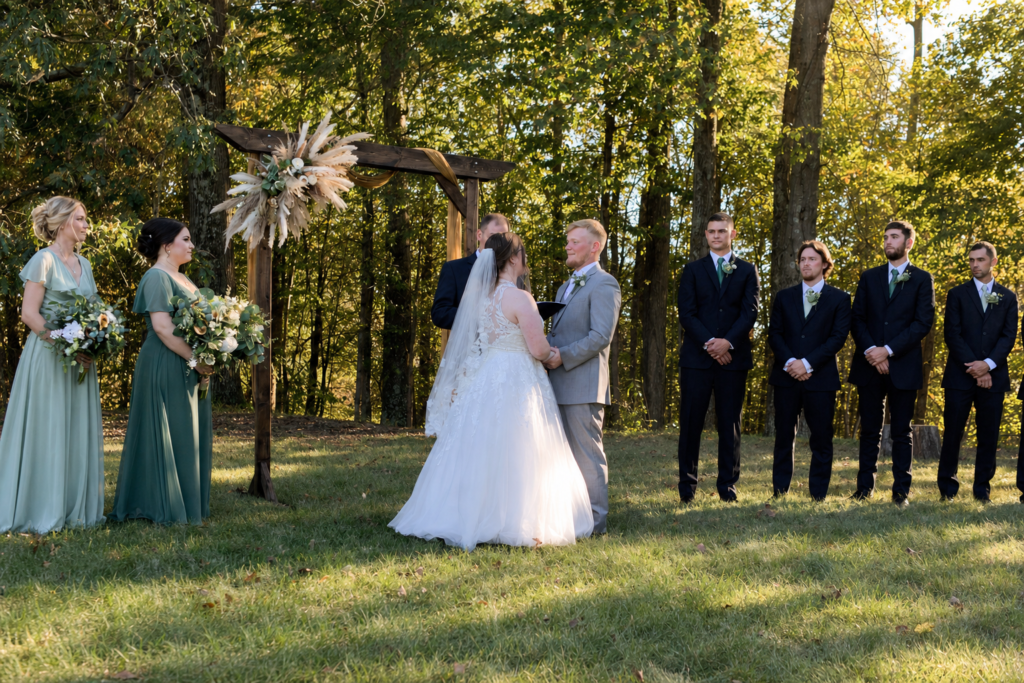 Bride and groom hold hands during an outdoor wedding ceremony under a wooden arch, flanked by bridesmaids and groomsmen