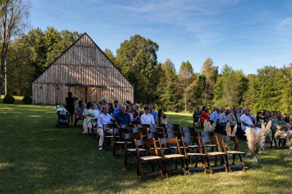 Guests seated in wooden chairs on a grassy lawn in front of a large wooden barn