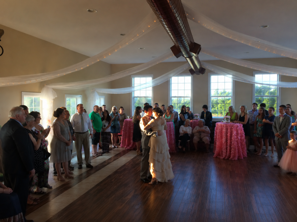 A bride and groom share a dance in a decorated reception hall surrounded by guests.