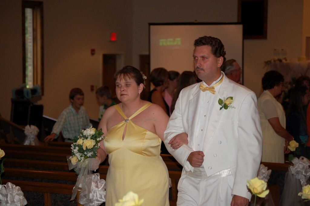 Man in a white tuxedo and woman in a yellow gown walk arm-in-arm with a bouquet of yellow and white roses.