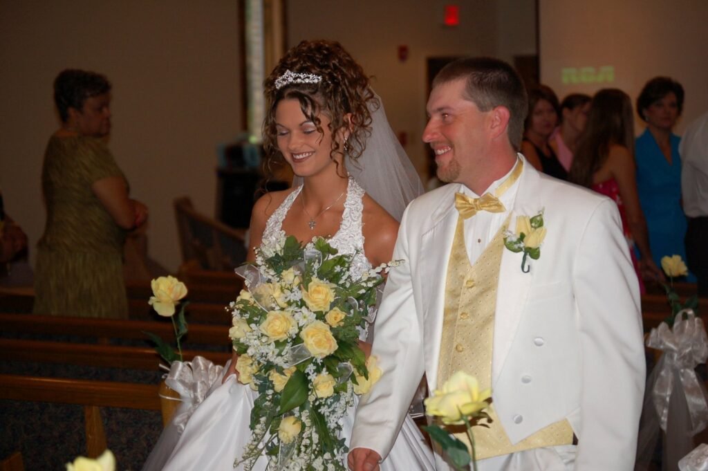 Smiling bride and groom in white wedding attire with yellow accents and a large yellow rose bouquet.