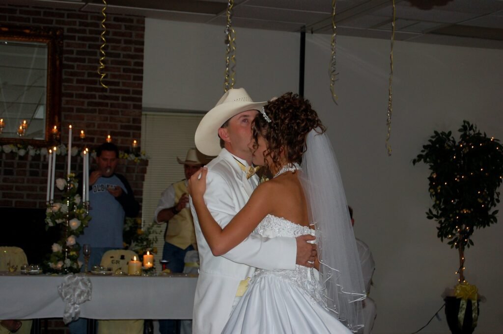 Bride in a white gown and veil kisses a groom in a white suit and cowboy hat at a wedding reception.