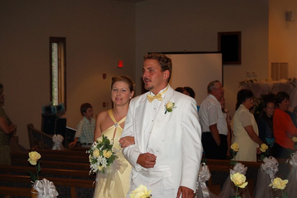A woman in a yellow gown and a man in a white tuxedo with a yellow bow tie stand together in a church.