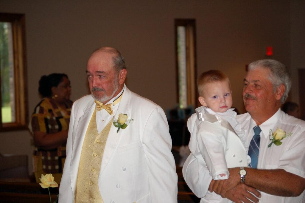 Three people in formal white attire, including a man in a yellow vest and a man holding a toddler.