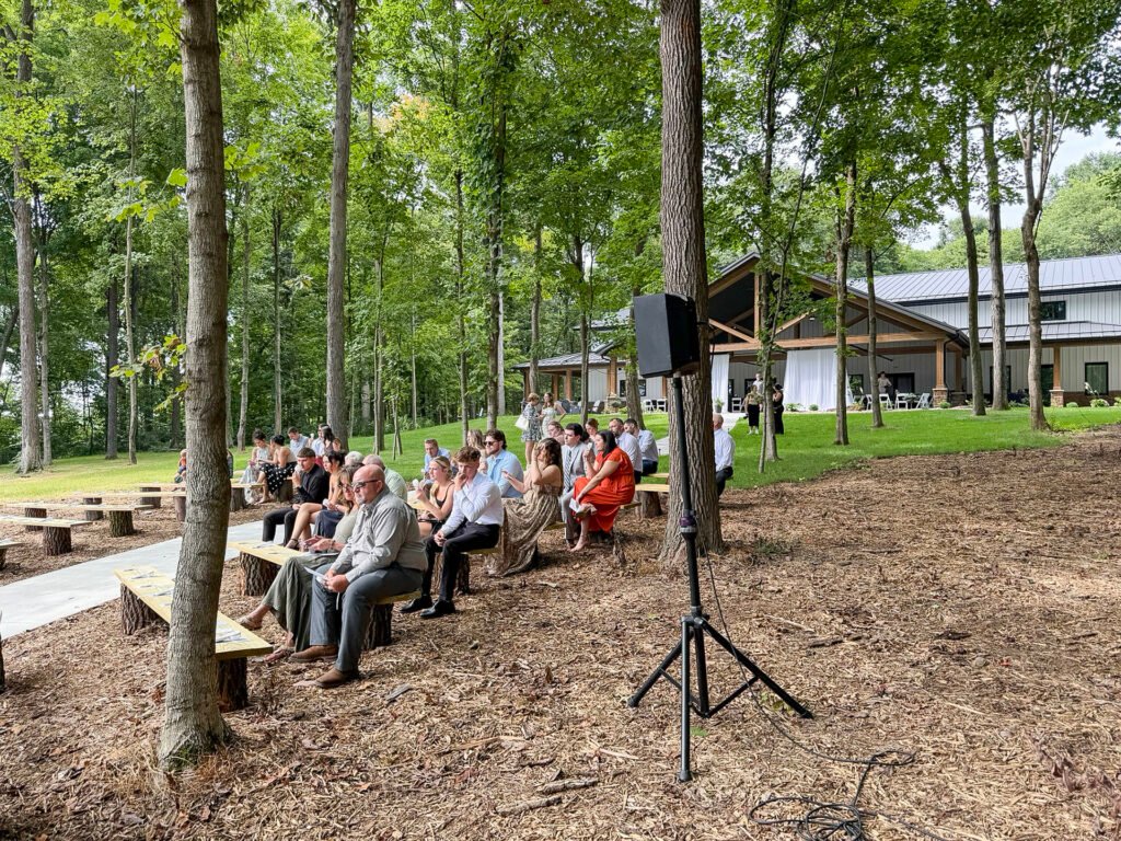 Guests seated on rustic wooden benches at the wooded outdoor ceremony site at Highbanks, with the venue building in the background.