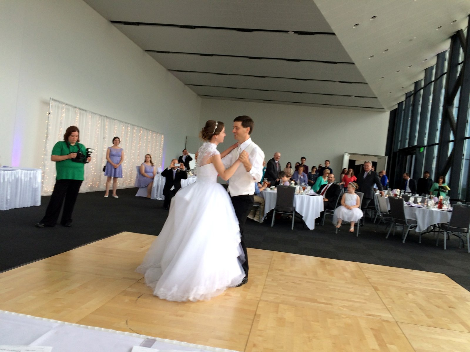 Bride and groom share their first dance on a wooden floor, surrounded by guests at a wedding reception.