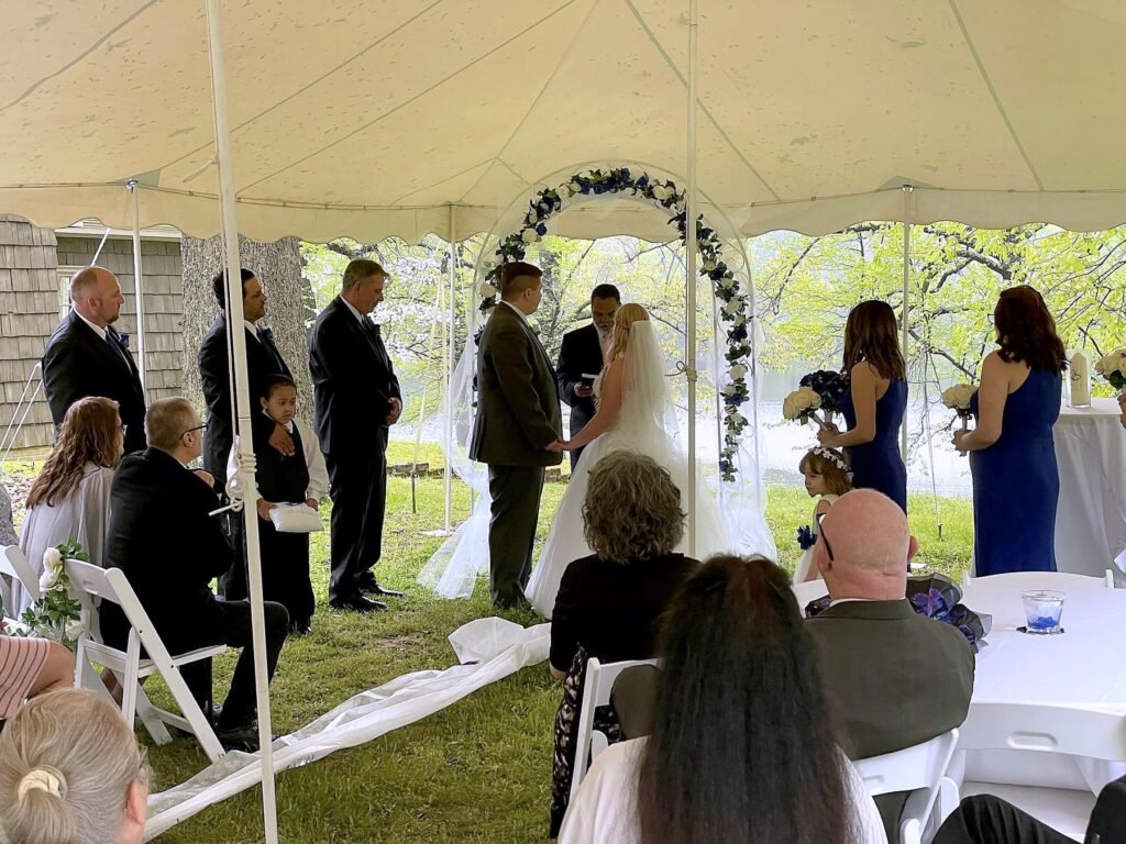 Madisonville Kentucky Wedding DJ 18 Wedding ceremony under a white tent with a bride and groom standing beneath a blue and white floral arch.